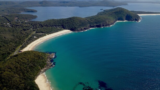 Une vue aérienne d’une zone côtière avec une plage de sable, des eaux turquoise et limpides, et des collines verdoyantes.