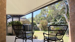 Een overdekt terras met twee zwarte metalen stoelen en een kleine tafel, met uitzicht op een tuin met bomen en een hek.