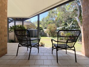 A covered patio with two black metal chairs and a small table, overlooking a garden with trees and a fence. - Aluna Holidays @ Boomerang - walk across the road to Boomerang Beach (north end) (Boomerang Beach)