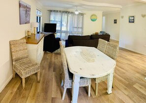 A dining area with a white table and wicker chairs, a black sofa, and a television. - Aluna Holidays @ Boomerang - walk across the road to Boomerang Beach (north end) (Boomerang Beach)
