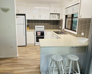 A modern kitchen with white cabinets, a microwave, a stove, and a sink. There are two stools at the island. - Aluna Holidays @ Boomerang - walk across the road to Boomerang Beach (north end) (Boomerang Beach)