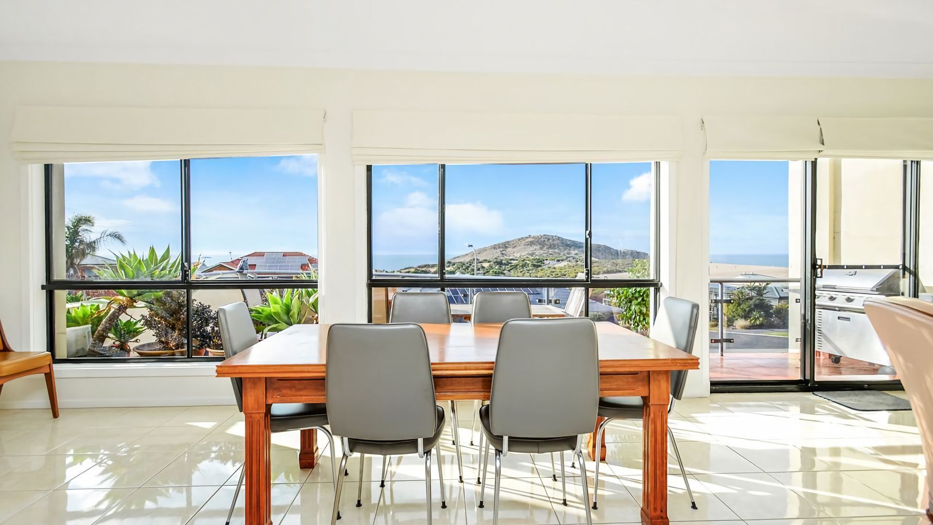 Un'ampia sala da pranzo con un grande tavolo e sedie, ampie finestre con vista su un paesaggio montuoso e sul mare, e un balcone con una griglia.