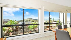 A modern room with large windows offering a view of a beach and mountains. There is a dining area with a table and chairs.