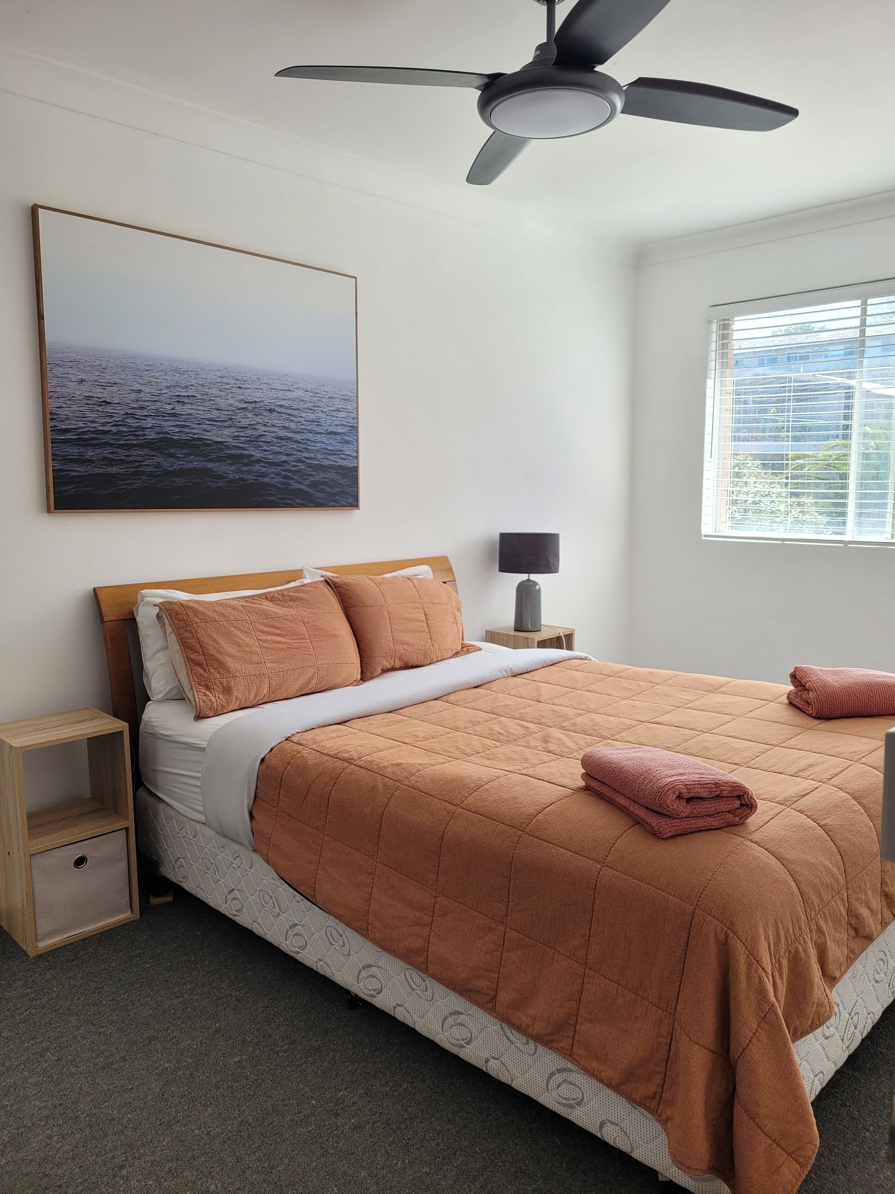 Une chambre à coucher avec un lit, un ventilateur de plafond, une table de chevet, une lampe, une fenêtre et un tableau représentant la mer.