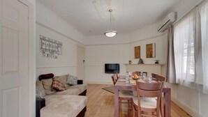 A modern living room with a sofa, dining table, and chairs. There is a flat-screen TV mounted on the wall.