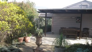 Un patio couvert avec une table et des chaises en bois, entouré de plantes en pot et d’un grand arbuste en fleurs.