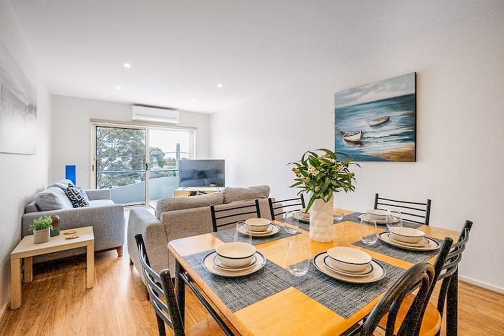 A dining area with a wooden table set for four, surrounded by chairs, with a plant centerpiece and a painting on the wall.