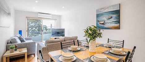 A dining area with a wooden table set for four, surrounded by chairs, with a plant centerpiece and a painting on the wall.