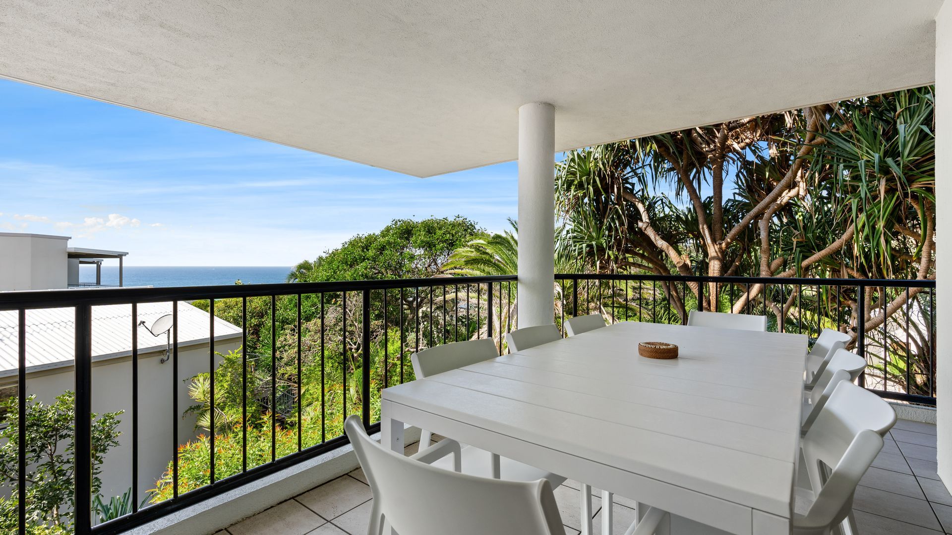 Un balcon avec une table et des chaises blanches, une vue sur l’océan et une végétation tropicale.