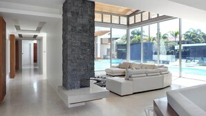 Un salon moderne avec une cheminée en pierre, un canapé d’angle blanc et une grande baie vitrée donnant sur une piscine.