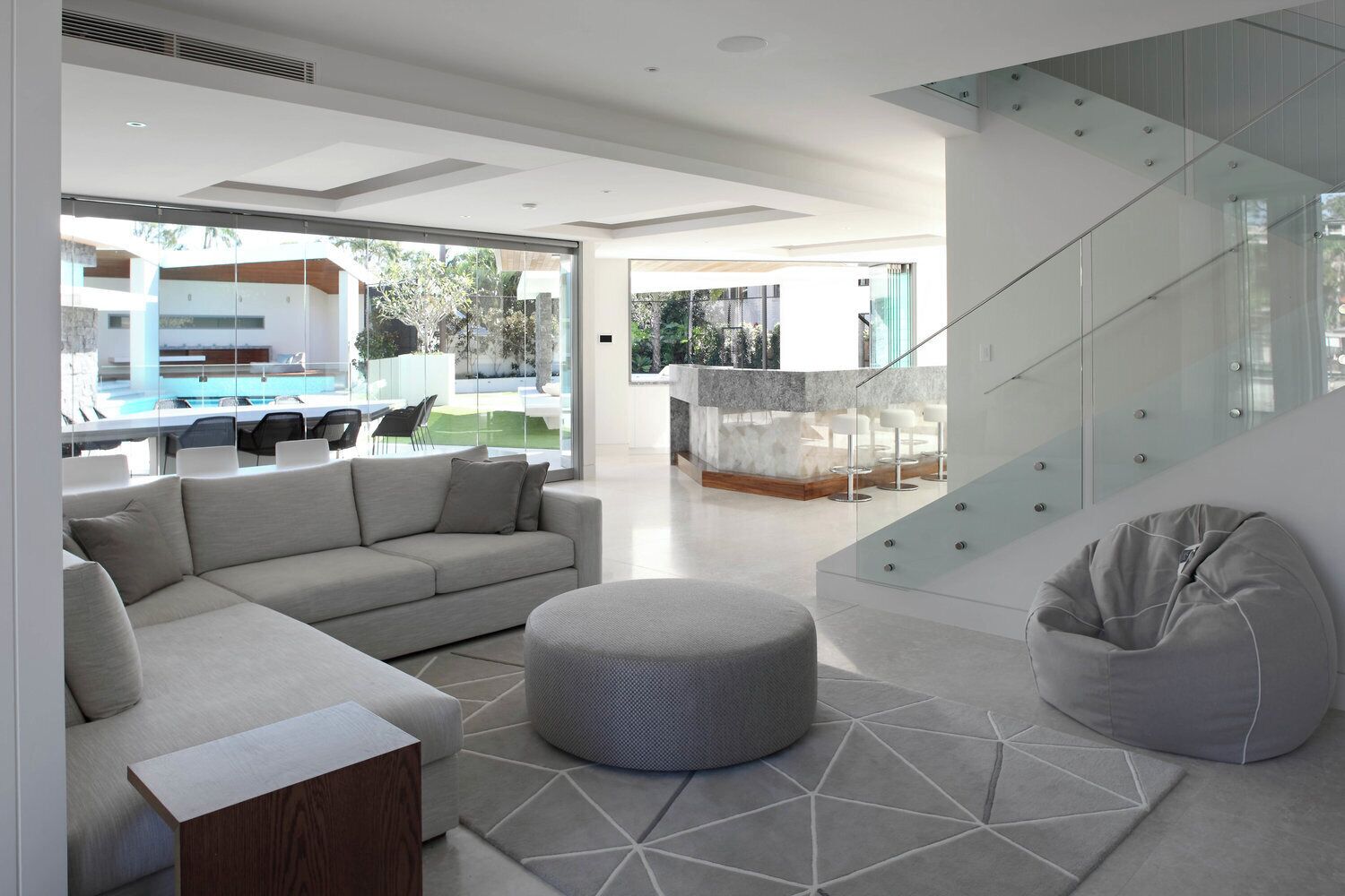 A modern living area with a sofa, ottoman, and bean bag chair, a glass staircase, and a pool view.