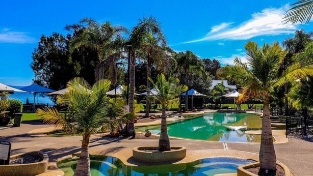 Zona de piscina con palmeras, sombrillas y cielo azul despejado.