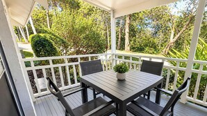 Un patio exterior cubierto con una mesa y sillas, rodeado por una barandilla blanca, con vista a un jardín con árboles y arbustos.