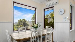 A dining room with a wooden table and white chairs, large windows with a view of trees and hills, and a wall-mounted clock. - Quingilli Luxury Chalet 3 Bedrooms (Ravensbourne)