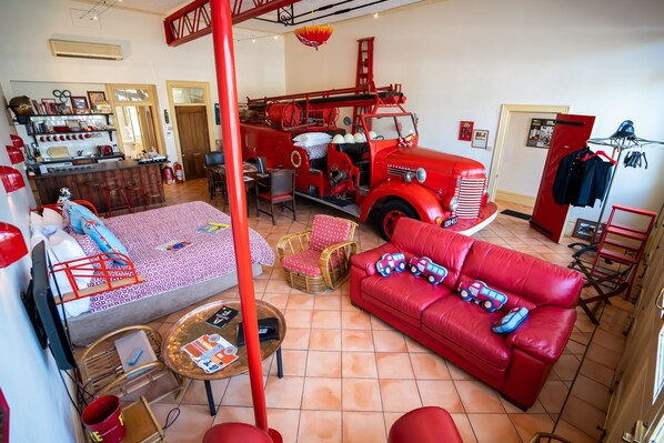 Una habitación con un camión de bomberos rojo, un sofá rojo, un comedor y una cama con colcha estampada.