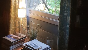 A room with a window covered by floral curtains, a desk with books and a lamp, and a view of greenery outside.