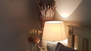 A bedroom with a bed, a bedside table with a lamp, decorative wall art, and a vase with dried flowers.