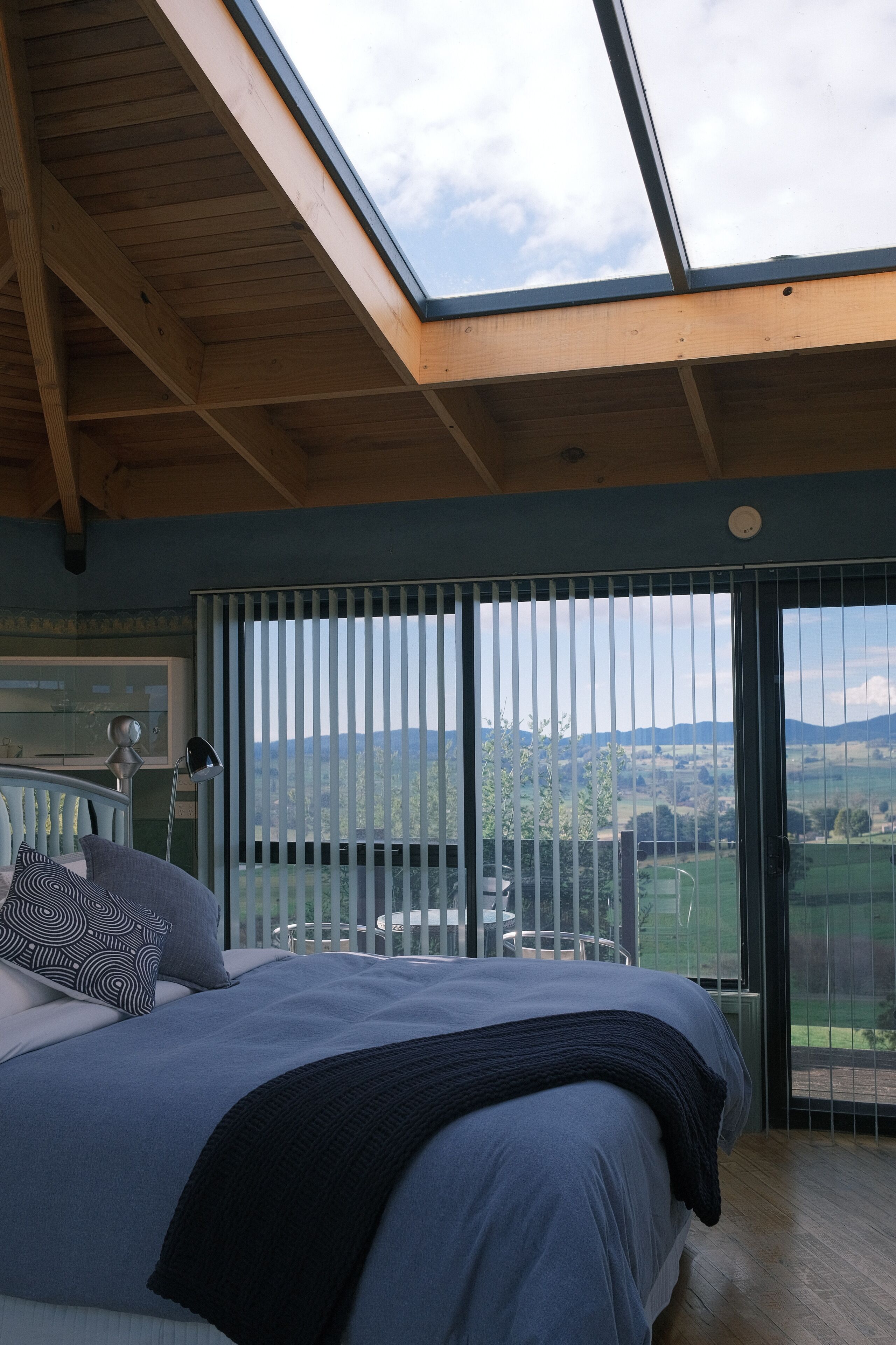Una camera da letto con un lucernario, un letto con copriletto blu, vista sulla campagna attraverso ampie finestre e un soffitto in legno.