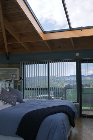 Un dormitorio con tragaluz, cama con cubrecama azul, vista al campo a través de amplias ventanas y techo de madera.