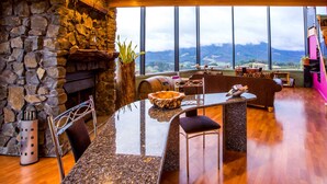 Un intérieur moderne avec une cheminée en pierre, un coin repas doté d’une table en verre et une grande fenêtre offrant une vue sur les montagnes.