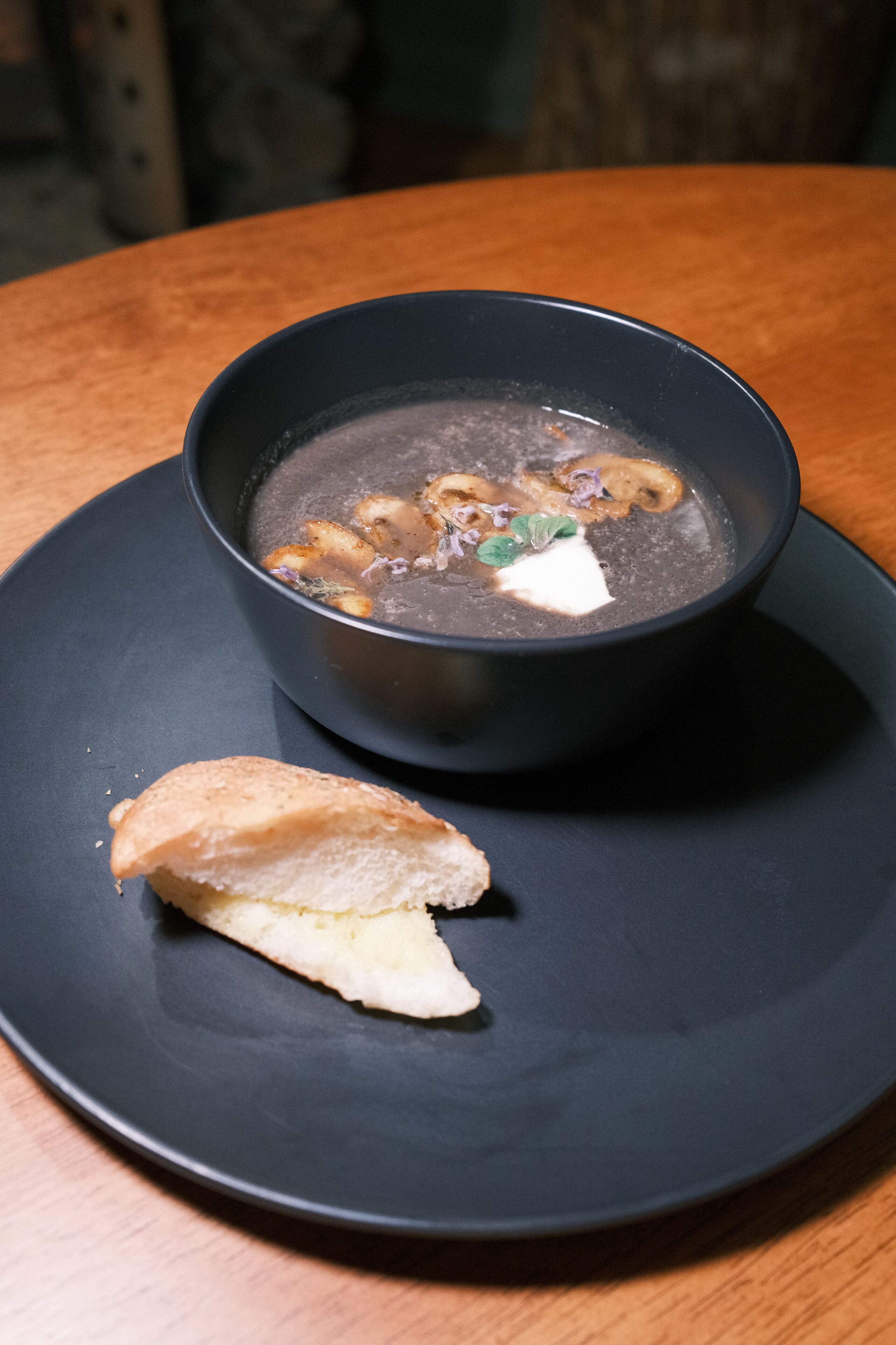 Un tazón oscuro de sopa con una rebanada de pan en un plato.
