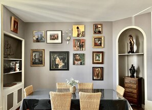A dining area with a table set for four, surrounded by wicker chairs. The walls are adorned with various framed artworks.