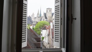 Una ventana con contraventanas abiertas, mostrando una vista del paisaje urbano con edificios y una aguja de iglesia.