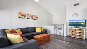 Een moderne woonkamer met een grijze bank, een houten salontafel en kleurrijke kussens. Er staat een flatscreen televisie op een witte dressoir en een trap die leidt naar een hoger niveau.