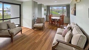 A spacious living area with a wooden floor, a sofa set, a dining table with chairs, and large windows offering a view of greenery. - Two Bedroom Apartment - Unit 4 Foreshores (Sunshine Coast)