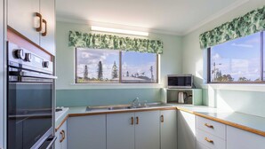 A kitchen with white cabinets, wooden handles, a microwave, and a window with a view of trees and a building. - Two Bedroom Apartment - Unit 4 Foreshores (Sunshine Coast)