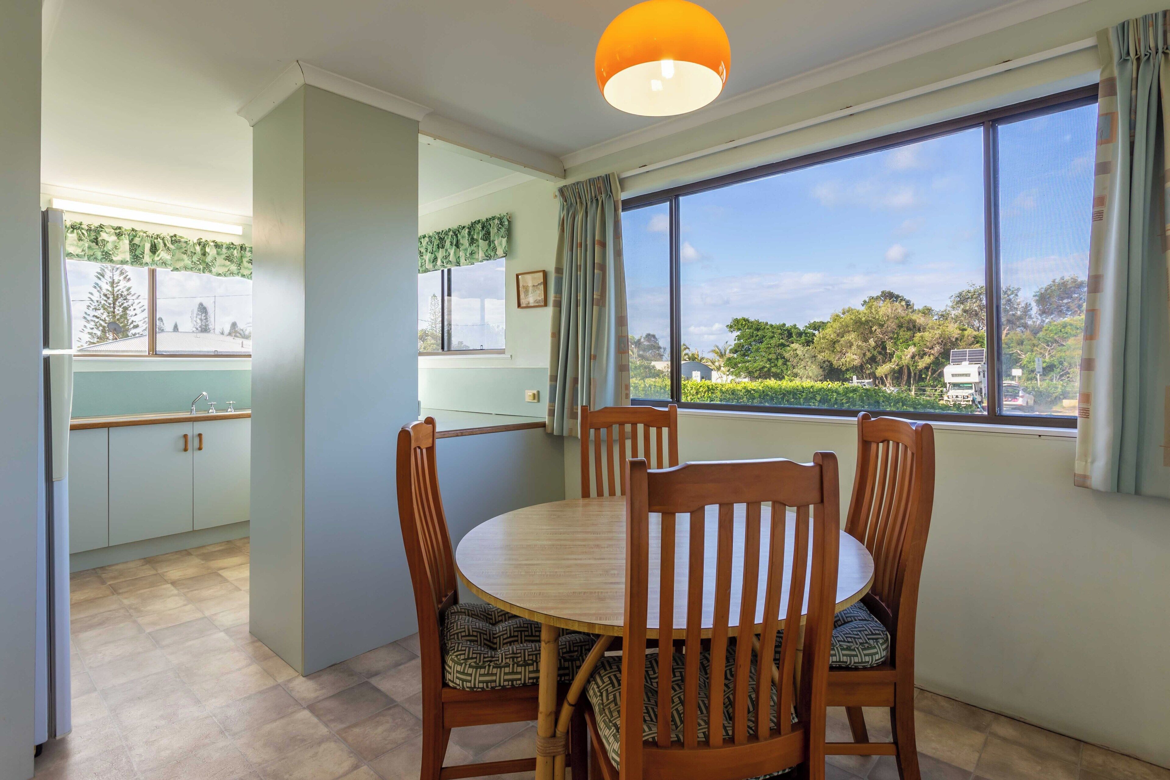 A dining area with a round table and four chairs, a window with a view of greenery and buildings, and a kitchen with cabinets and a window with curtains.