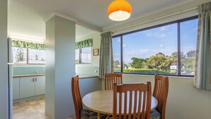 A dining area with a round table and four chairs, a window with a view of greenery and buildings, and a kitchen with cabinets and a window with curtains. - Two Bedroom Apartment - Unit 4 Foreshores (Sunshine Coast)