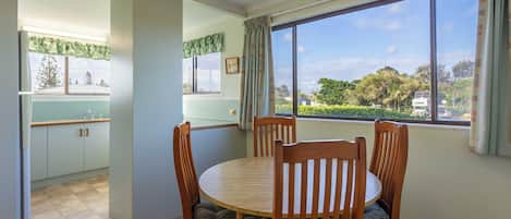 A dining area with a round table and four chairs, a window with a view of greenery and buildings, and a kitchen with cabinets and a window with curtains.