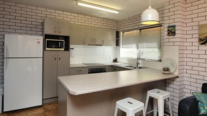 Une cuisine de taille réduite avec des meubles blancs, un four à micro-ondes, un évier et une fenêtre équipée de stores.