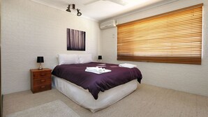 Une chambre à coucher comprenant un lit, deux tables de chevet, un climatiseur fixé au mur et une fenêtre avec des stores.
