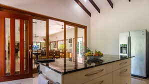 A modern kitchen with a marble countertop, a refrigerator, and a dining area visible through a glass door.
