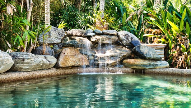 A clear pool with a waterfall feature surrounded by rocks and lush greenery.
