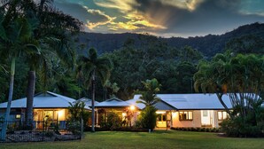 A two-story house with a well-maintained lawn, surrounded by tropical vegetation and a hilly backdrop.