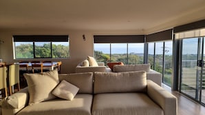Ein modernes Wohnzimmer mit einer großen Couch, einem Esstisch und großen Fenstern, die einen Ausblick ins Freie bieten.