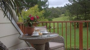 Une table à plateau en verre, sur laquelle reposent une tasse et une soucoupe blanches, un pot de fleurs avec des fleurs rouges, et une chaise en osier sur un balcon offrant une vue sur un paysage verdoyant.