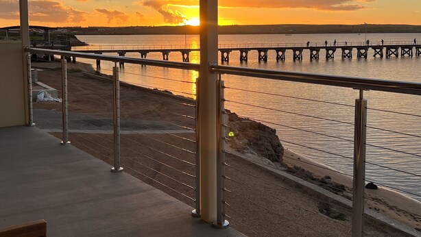 Ein Balkon mit Blick auf einen Sonnenuntergang über einem Gewässer, eine Mole, die sich ins Wasser erstreckt, und ein Sandstrand.