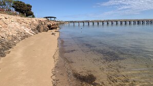 Ein Strand mit felsiger Küste, einer Pier, die sich über ruhiges Wasser erstreckt, und ein Gebäude auf der linken Seite.