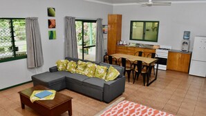 A living room with a sofa, a coffee table, and a patterned rug. There is a dining area with a table and chairs, and a kitchen with cabinets and a refrigerator. - Tuteveras' Arms - Arorangi (Arorangi)