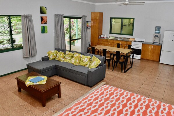 A living room with a sofa, a coffee table, and a patterned rug. There is a dining area with a table and chairs, and a kitchen with cabinets and a refrigerator.