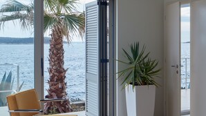 A modern living room with a view of the sea, featuring a sofa, armchair, ottoman, and a potted plant. - A Place Where The Garden Ends And The Sea Begins (Milna)
