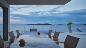 A modern dining area with a glass table, chairs, and a view of the sea. - A Place Where The Garden Ends And The Sea Begins (Milna)