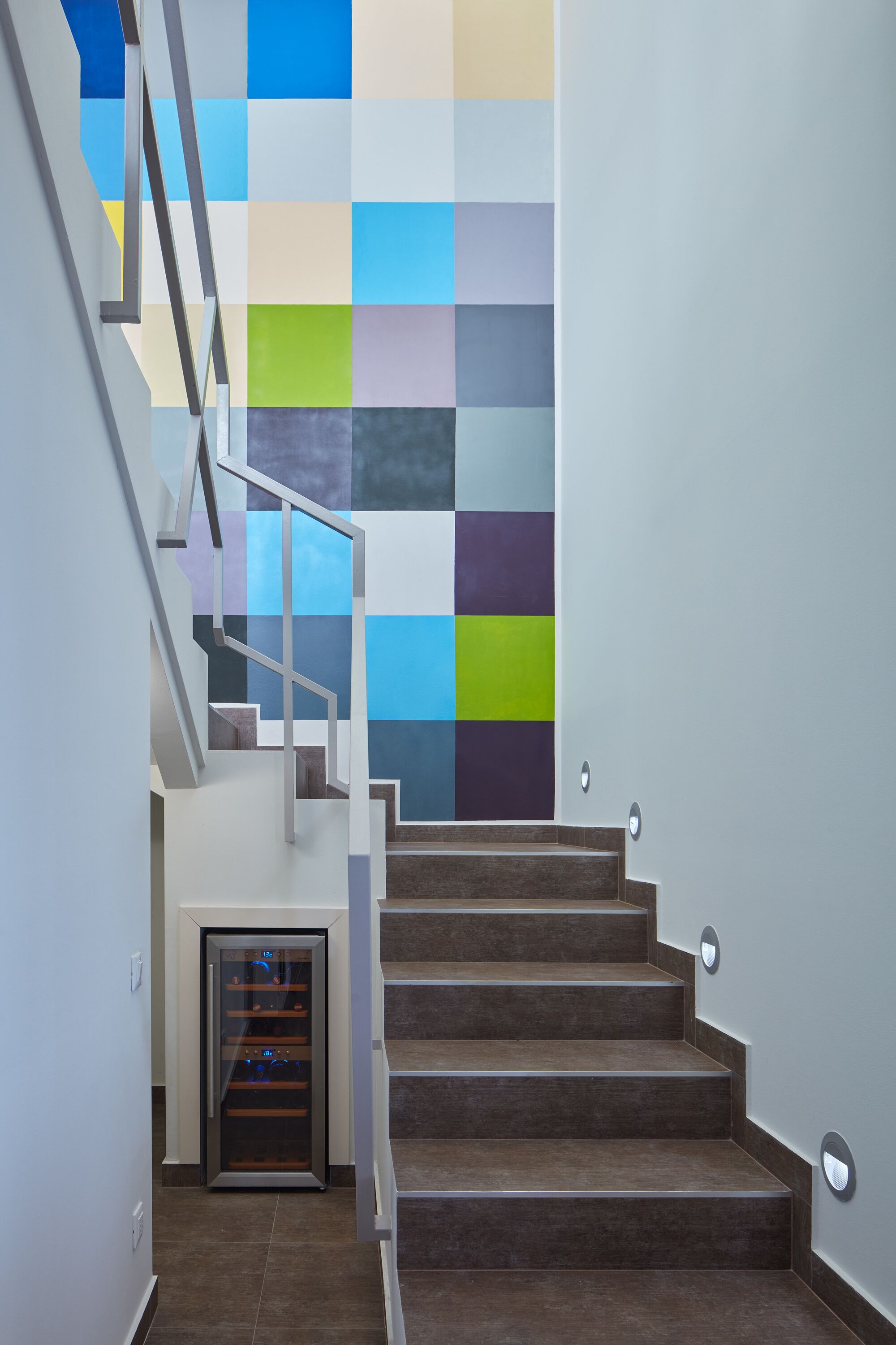 A staircase with a modern design, a glass door at the bottom, and a wall with a colorful square pattern.