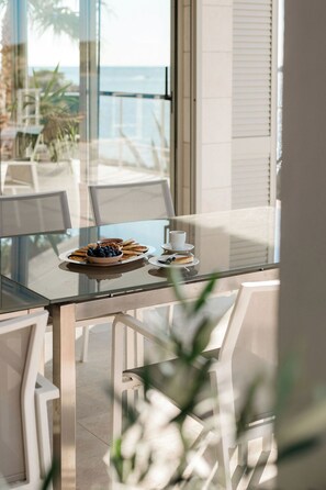 A dining area with a glass table, white chairs, and a view of the ocean. - A Place Where The Garden Ends And The Sea Begins (Milna)