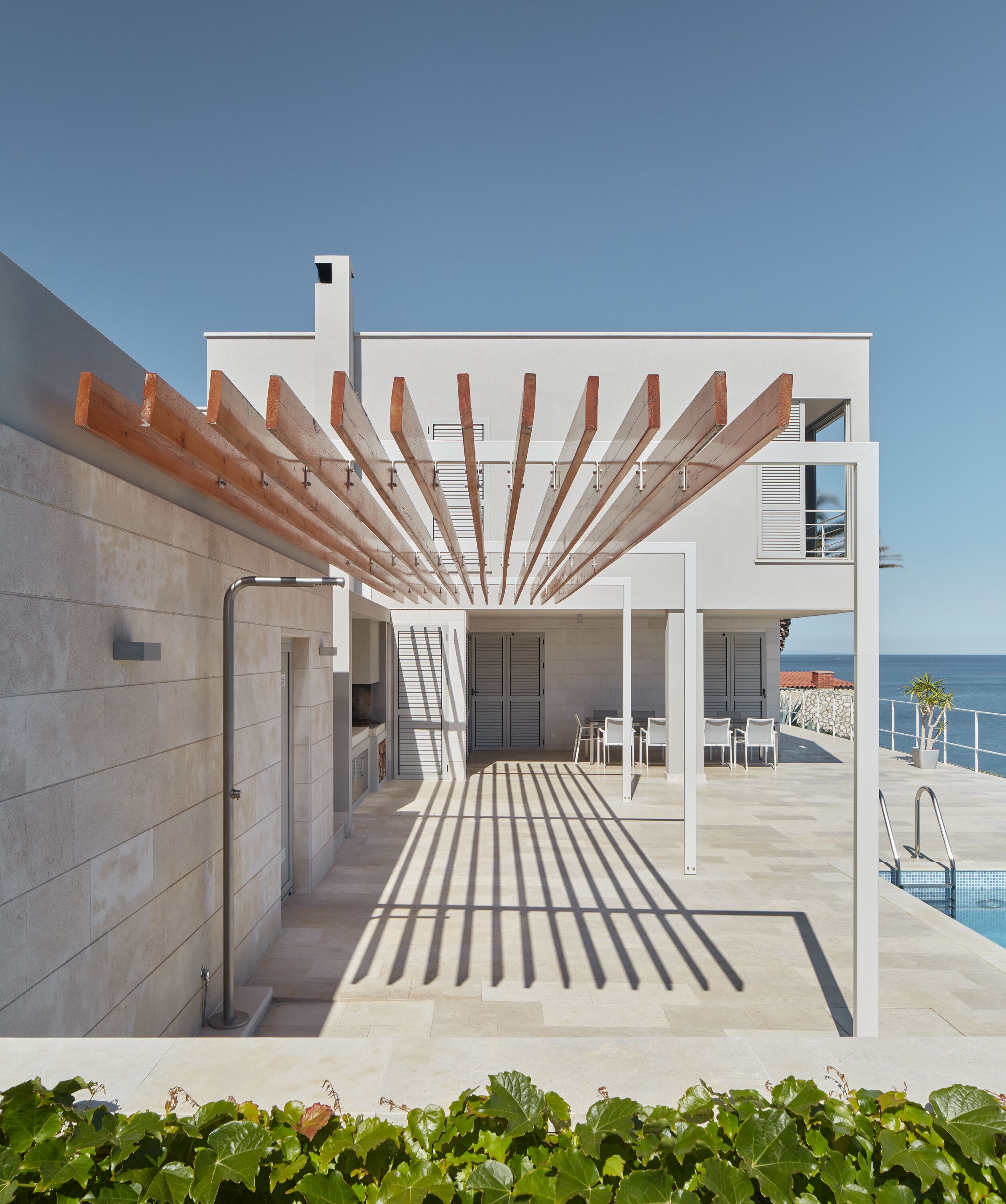 Un patio exterior moderno con pérgola, piscina y vista al mar.