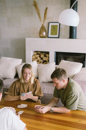 A group of people playing cards at a wooden table in a living room with a fireplace, cushions, and a vase with dried plants. - A Place Where The Garden Ends And The Sea Begins (Milna)